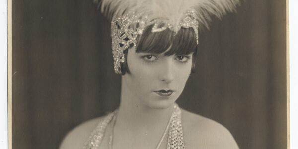 Photo of Louise Brooks in medium shot, wearing a white feathered headdress, looking at camera