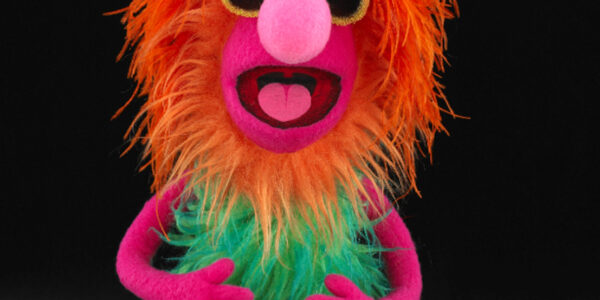 Puppet with fiery-red fur on its head, pink nose, and green torso, mouth open and looking at camera, wearing sunglasses
