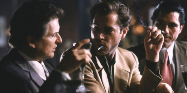 Two tough-looking gangsters sit at a bar, the one on right with a cigarette in his mouth