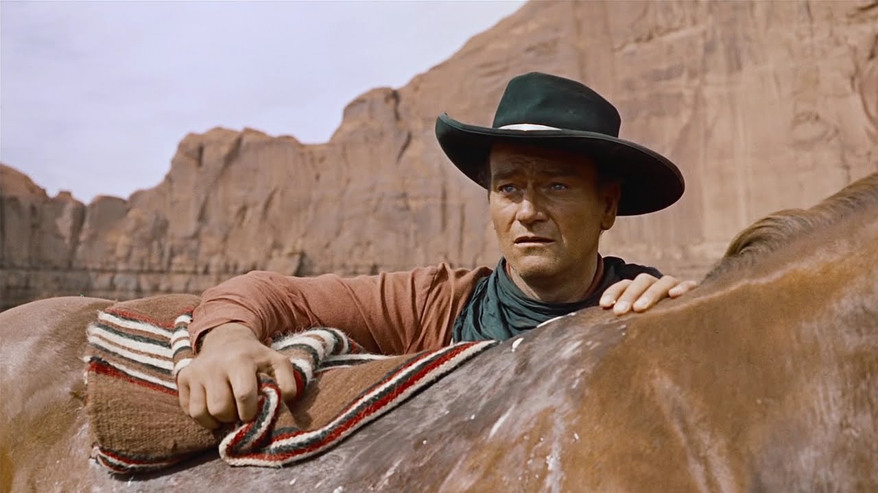 A man holds a horse's saddle as he stands next to the horse in front of a beige canyon