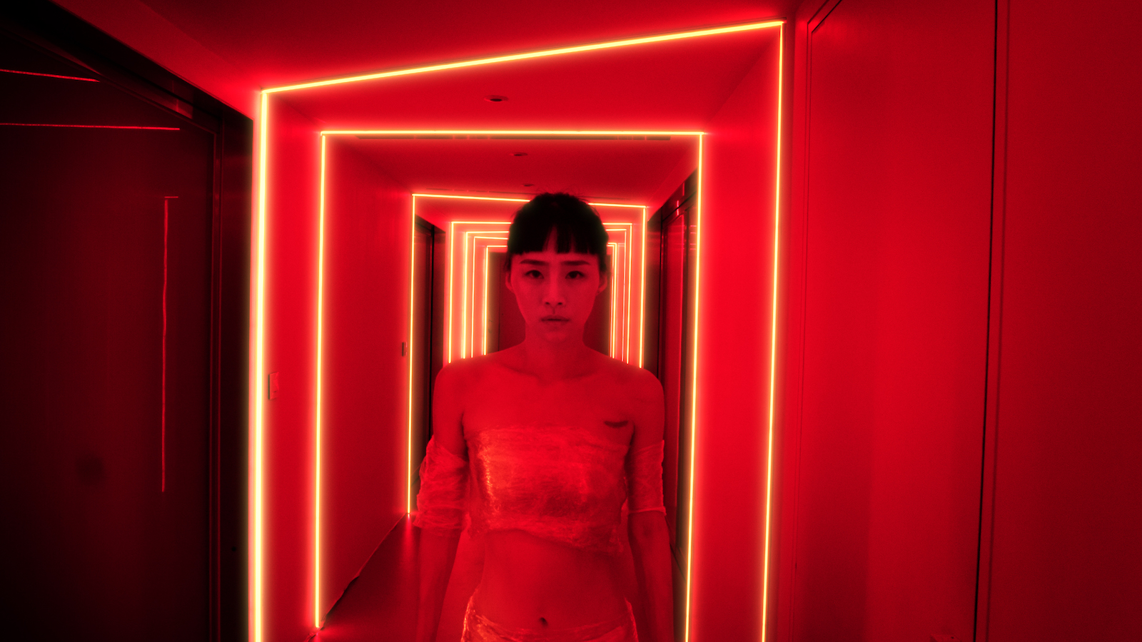 A woman stands in a dark, red-drenched hallway, looking at camera intensely.