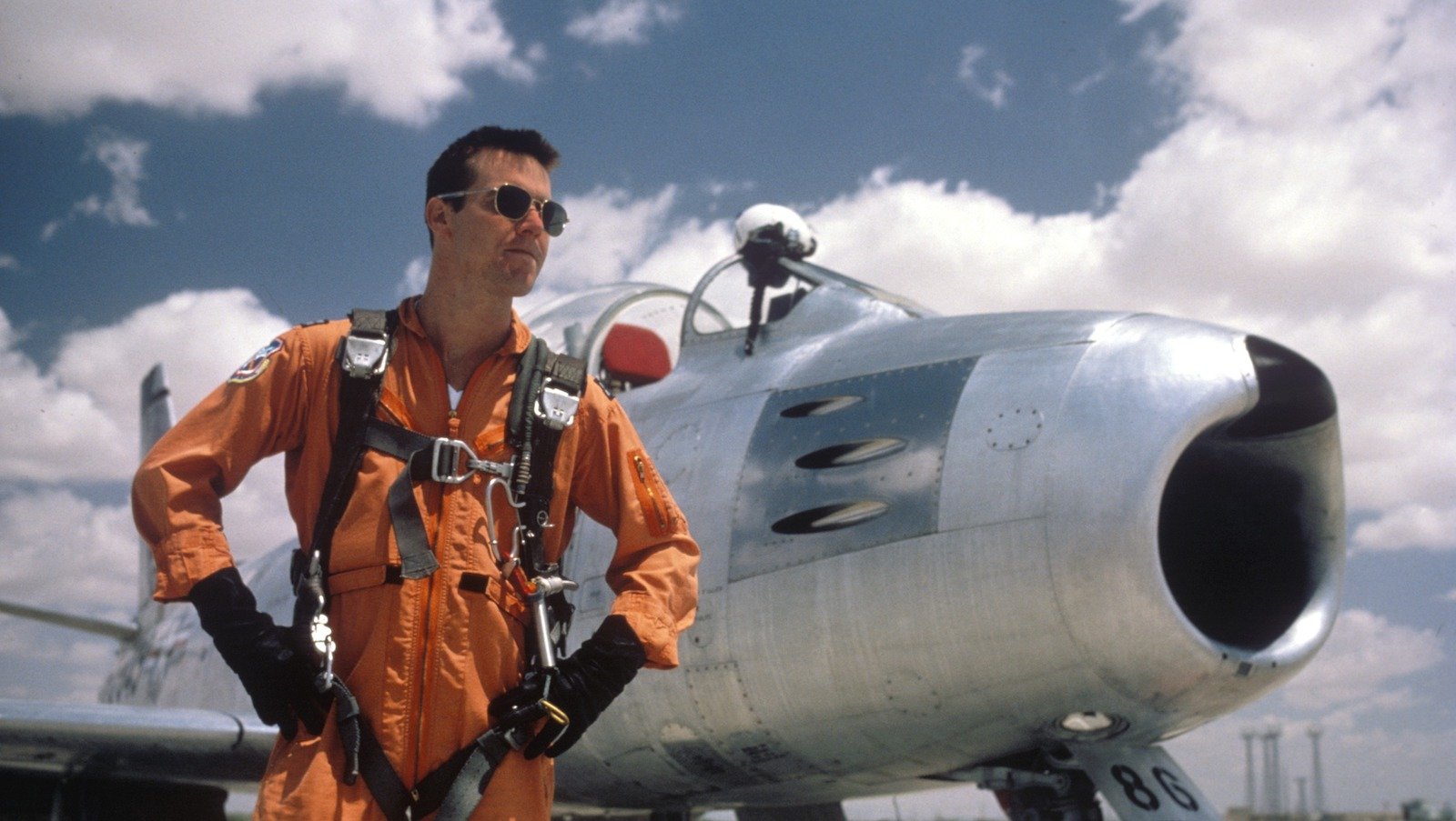 A test pilot in orange and aviator sunglasses standing in front of an airplane
