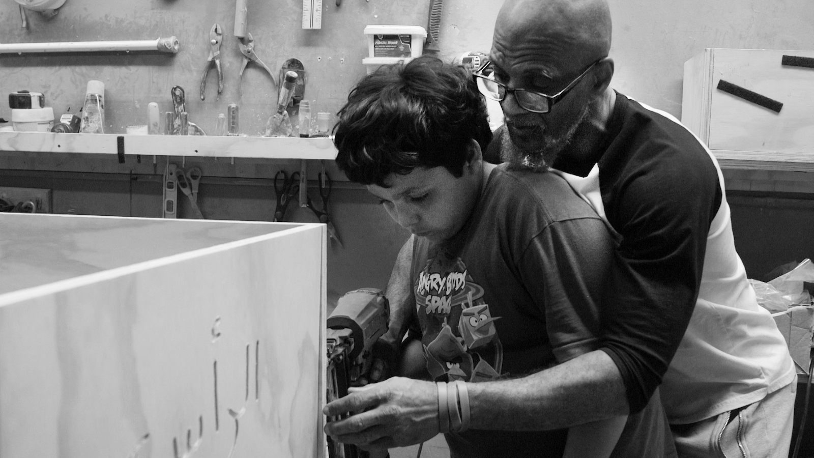An older man has his arms around a young boy as he teaches him carpentry