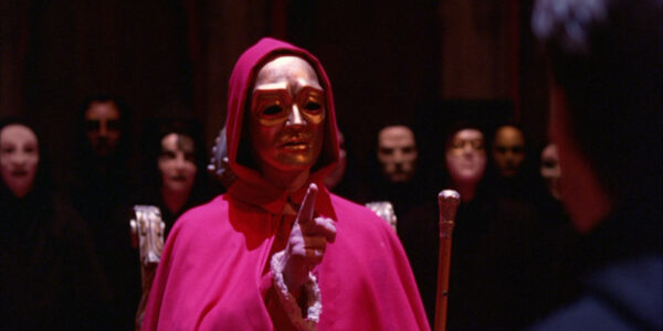 An ominous figure in a red cloak and gold mask points at the camera.