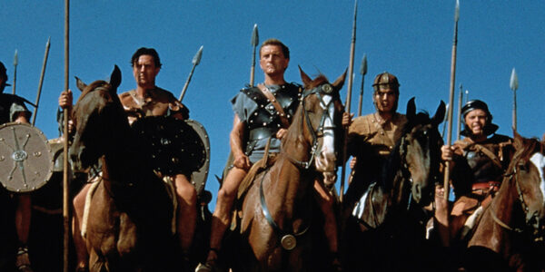 Spartacus and his fellow gladiators lined up on horses and carrying spears against a blue sky