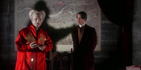 The ancient vampire Dracula dressed in a red robe in the foreground, while his shadow on the wall mimics strangling the young man standing behind him.