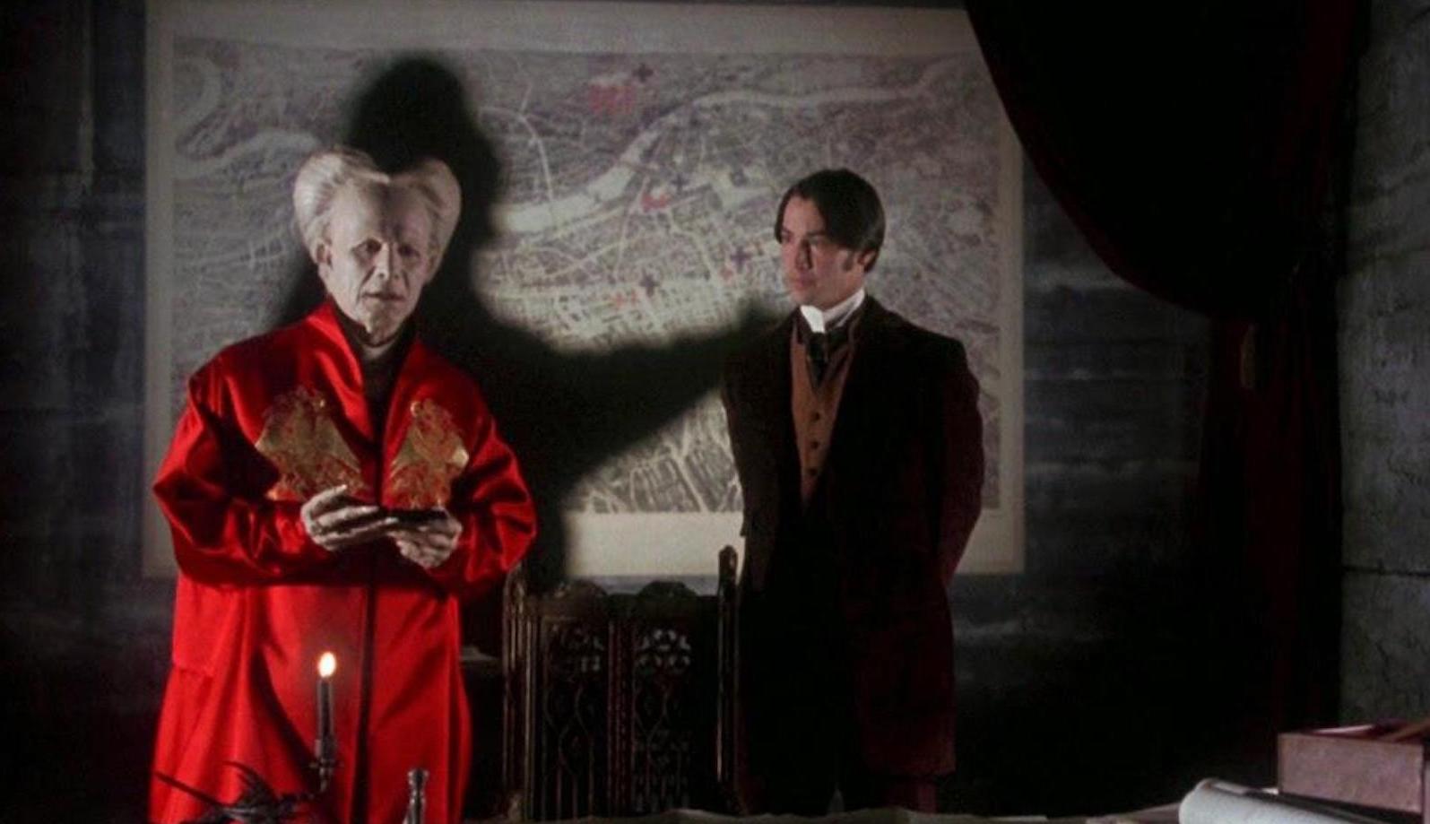 The ancient vampire Dracula dressed in a red robe in the foreground, while his shadow on the wall mimics strangling the young man standing behind him.