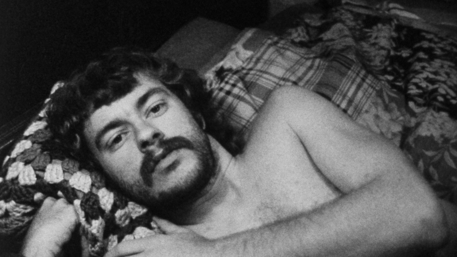 A bearded, shirtless man reclines in bed and looks at camera.