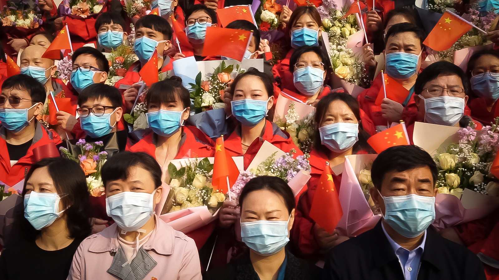 A crowd of face-masked people in China look towards the camera, many holding flowers and wearing red.