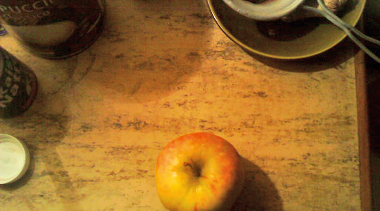 A photographic still life of an apple on a table, with mug and saucer nearby.