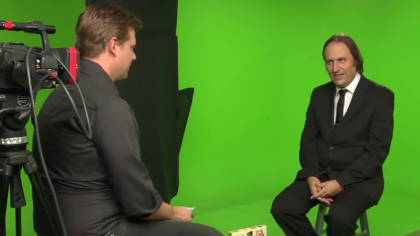 Two men sit on stools against a green-screen background