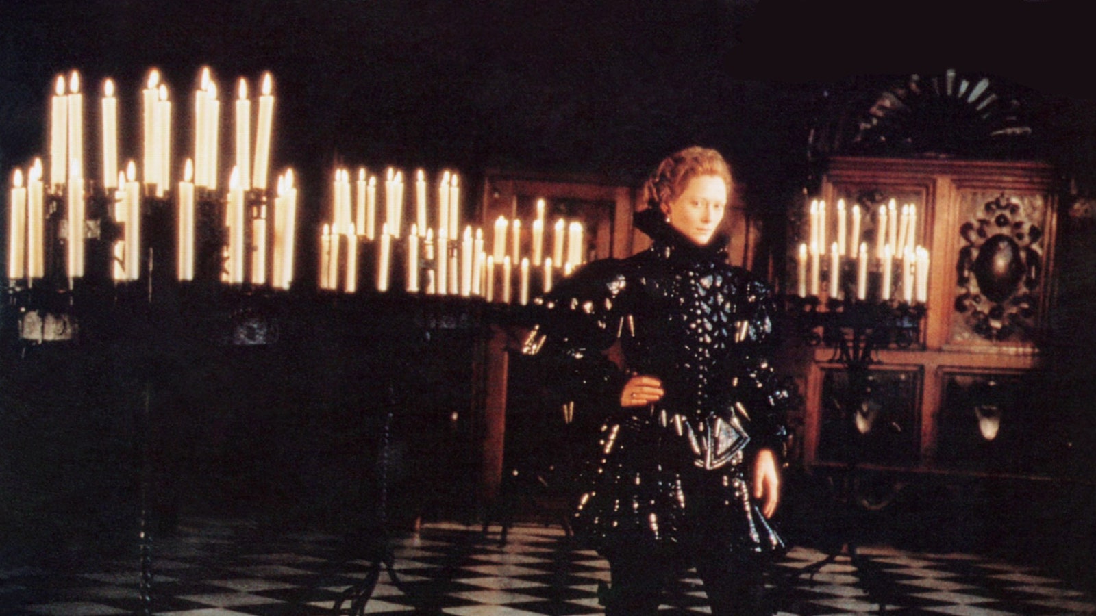 An androgynous English poet of the Elizabethan era stands in a dark room full of lit candles.