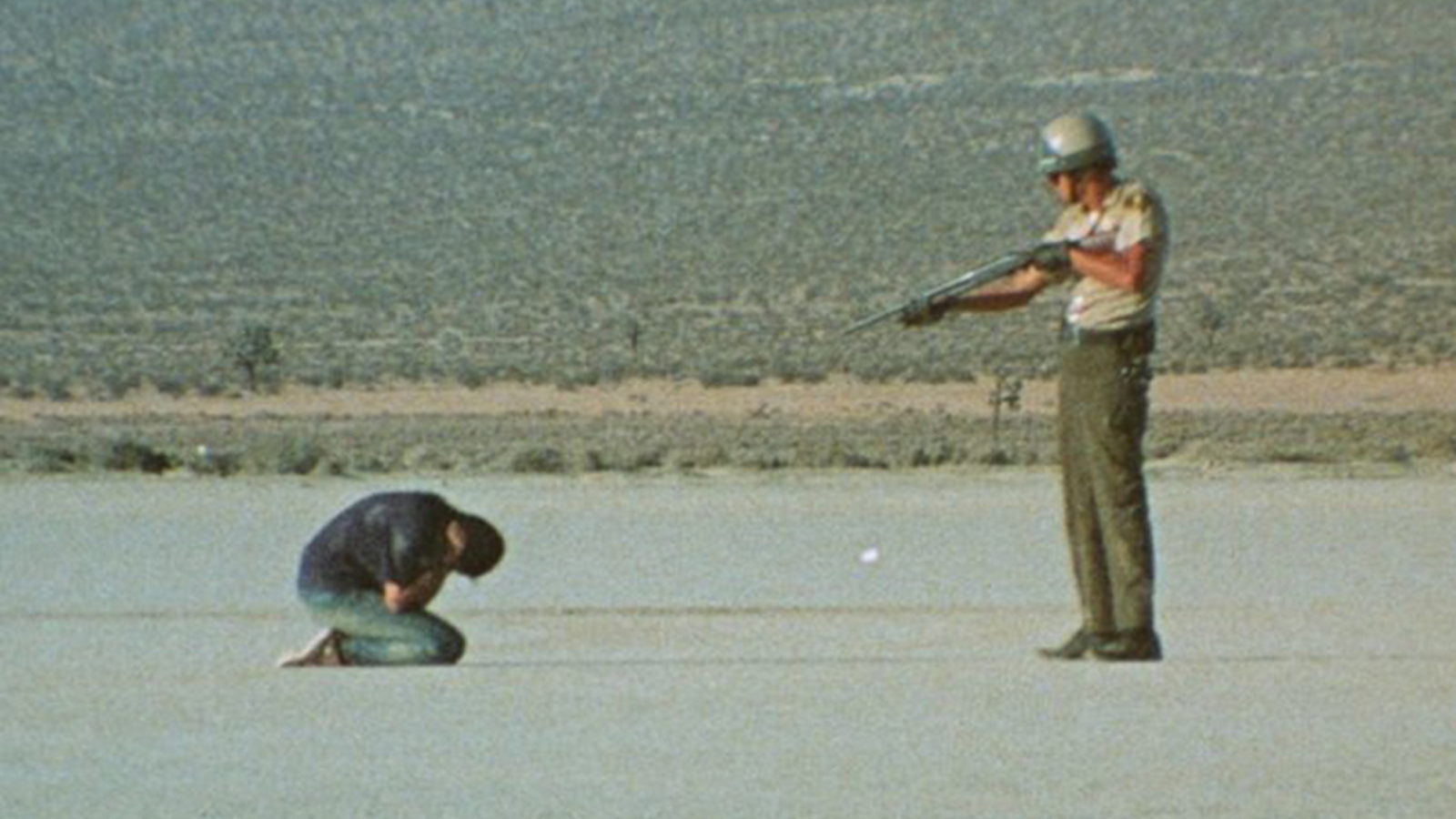 In a vast desert landscape, a policeman aims a rifle at a man cowering before him.