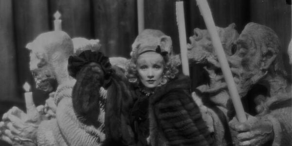 The actress Marlene Dietrich in a thick fur coat and hat looking at camera, surrounded by elaborate candelabras in the shape of gargoyles
