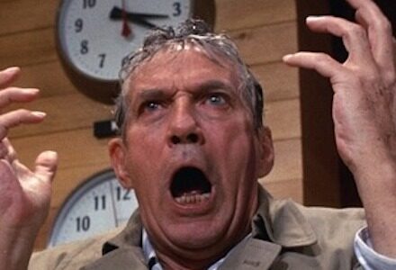 A man in a trenchcoat, wet from the rain, raises his hands and screams in front of a clock that reads 3:30.