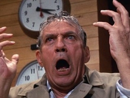 A man in a trenchcoat, wet from the rain, raises his hands and screams in front of a clock that reads 3:30.
