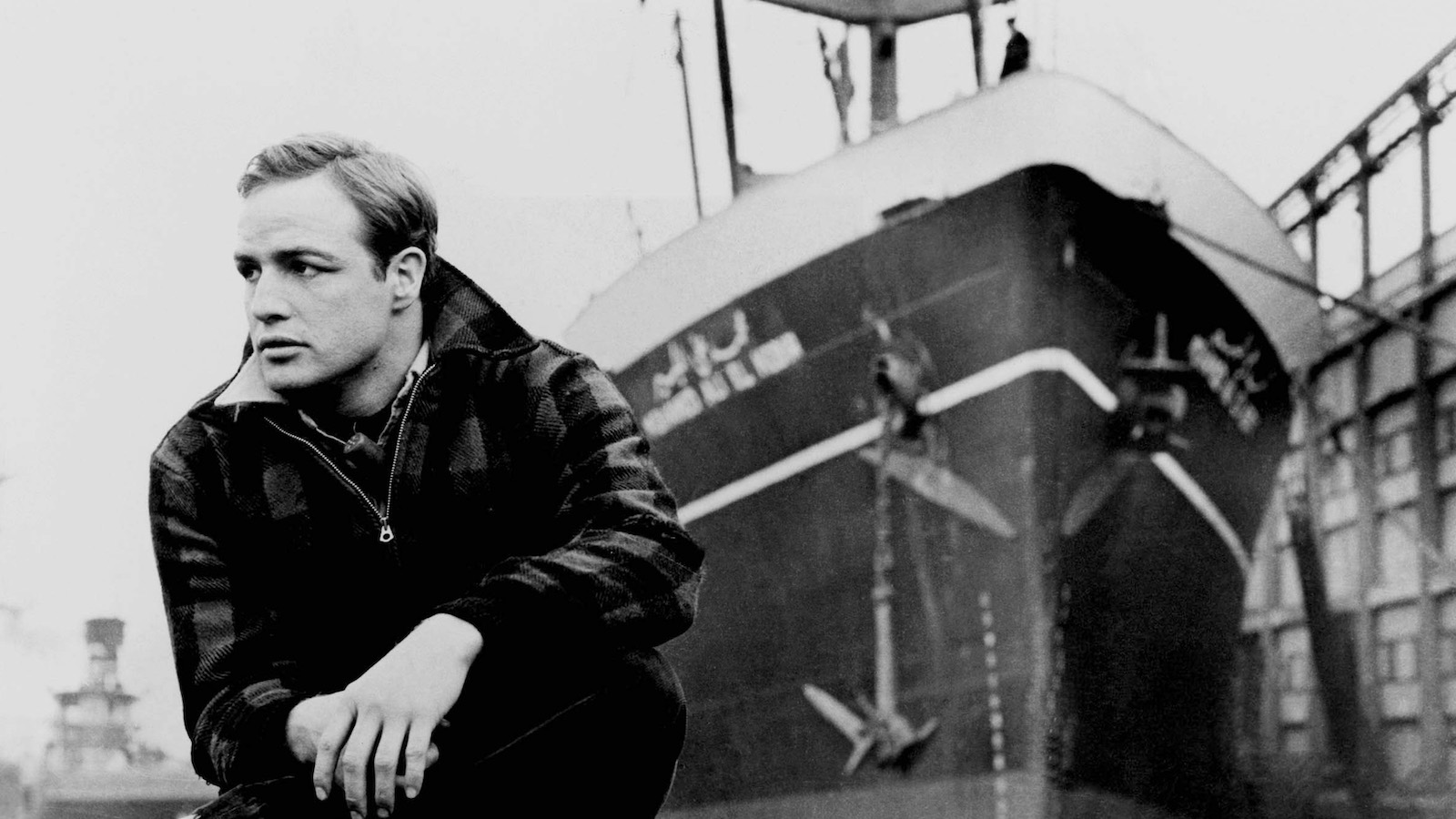 A man crouches by a dockside pier, a large ship behind him in the background