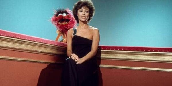 A woman in a black evening dress sits in the corner of a set, with a red Muppet behind her.