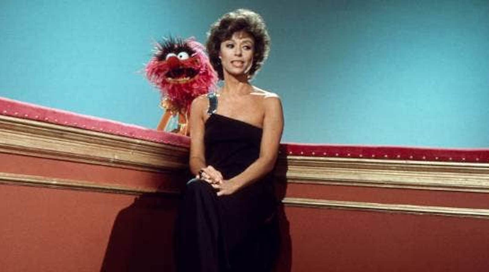 A woman in a black evening dress sits in the corner of a set, with a red Muppet behind her.