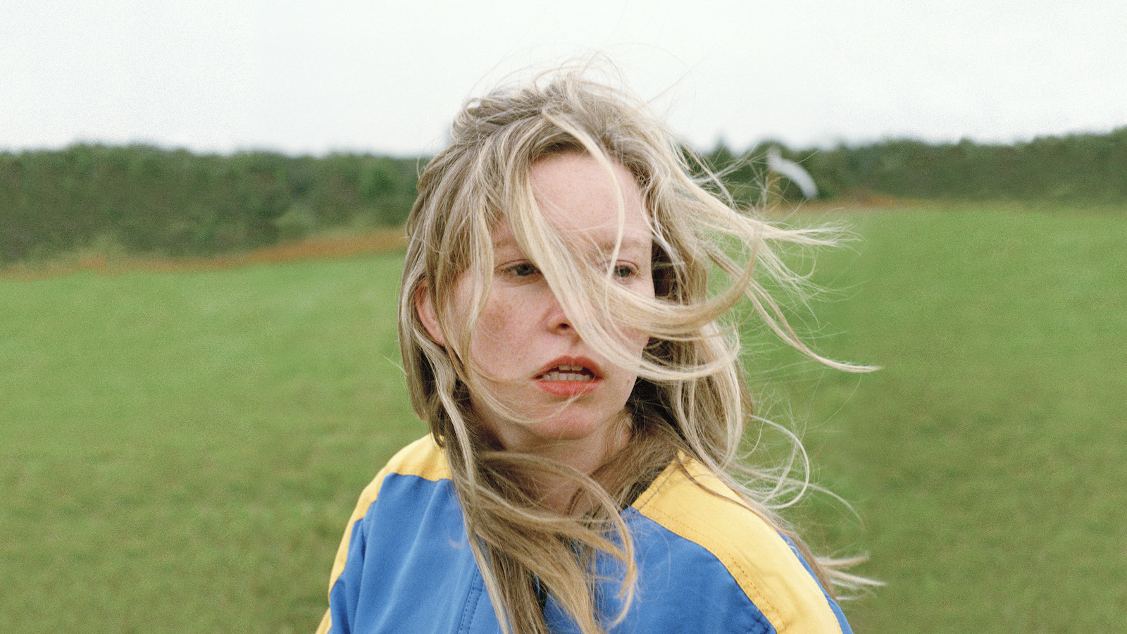 A distressed young woman stands in a field with long windswept blonde hair in her face.