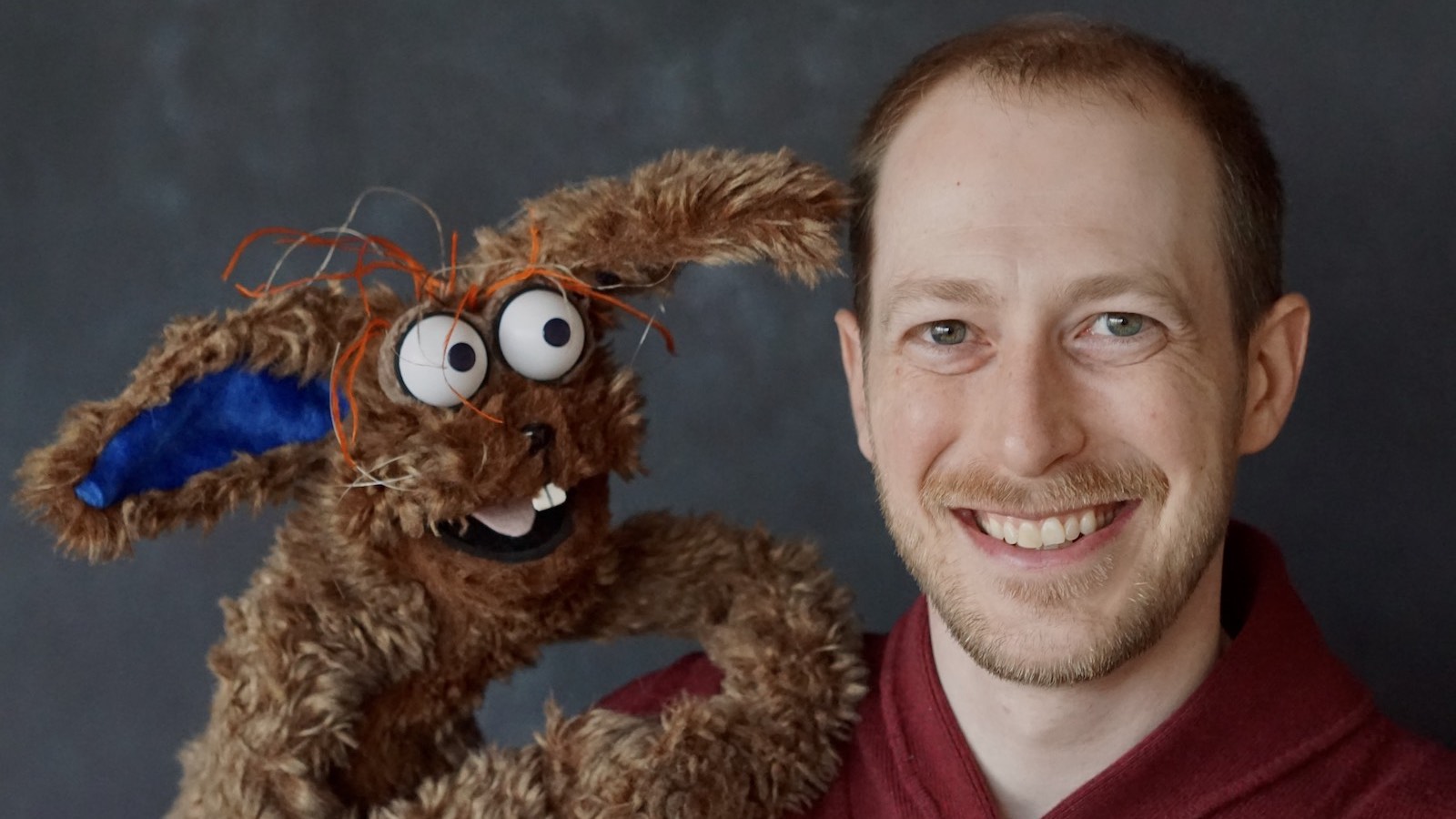 Puppeteer Brian Carson smiles at camera in closeup next to his puppet, the brown rabbit Silly Willy looking at him