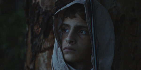 A young melancholy boy, his face draped by a hood, looks off-camera.