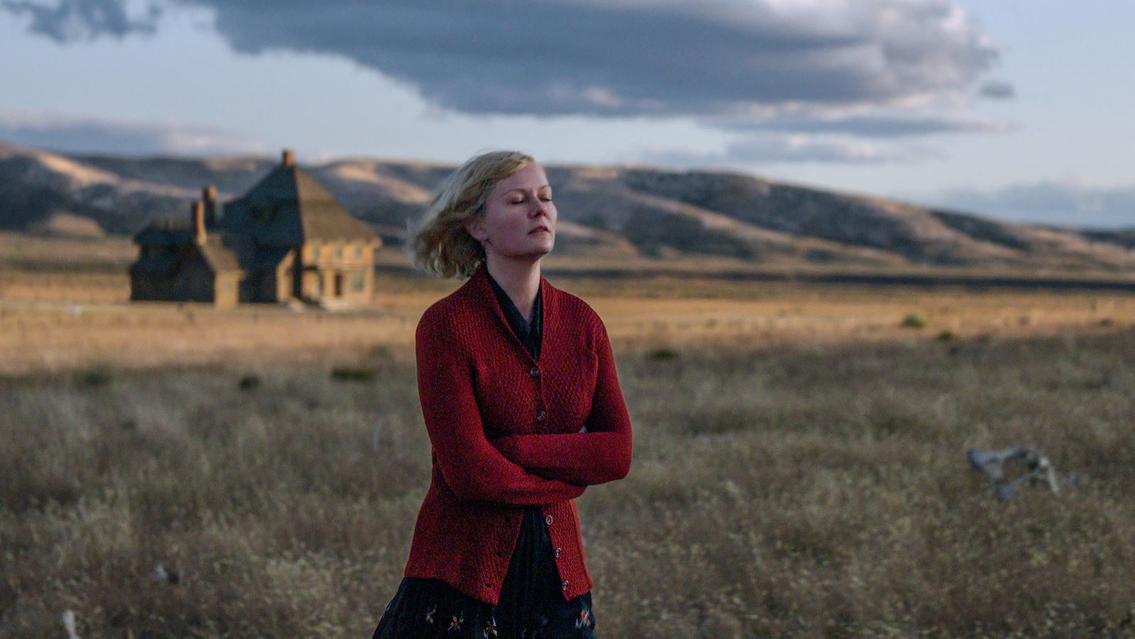 A woman (Kirsten Dunst) in a red sweater stands with her arms crossed against a vast Montana mountain landscape.