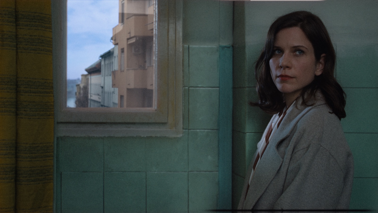 A woman with long dark hair looks at the camera standing next to a window looking out onto a city.