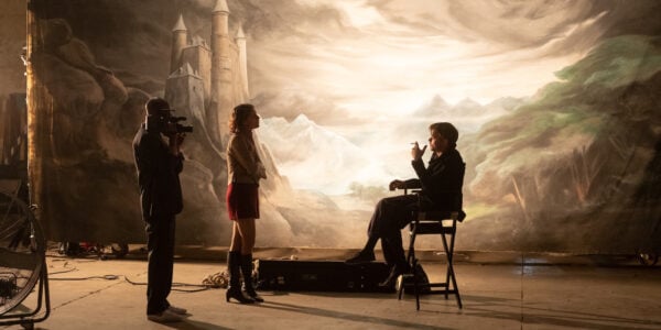 Three people in silhouette, one sitting in a director's chair, on a movie set.