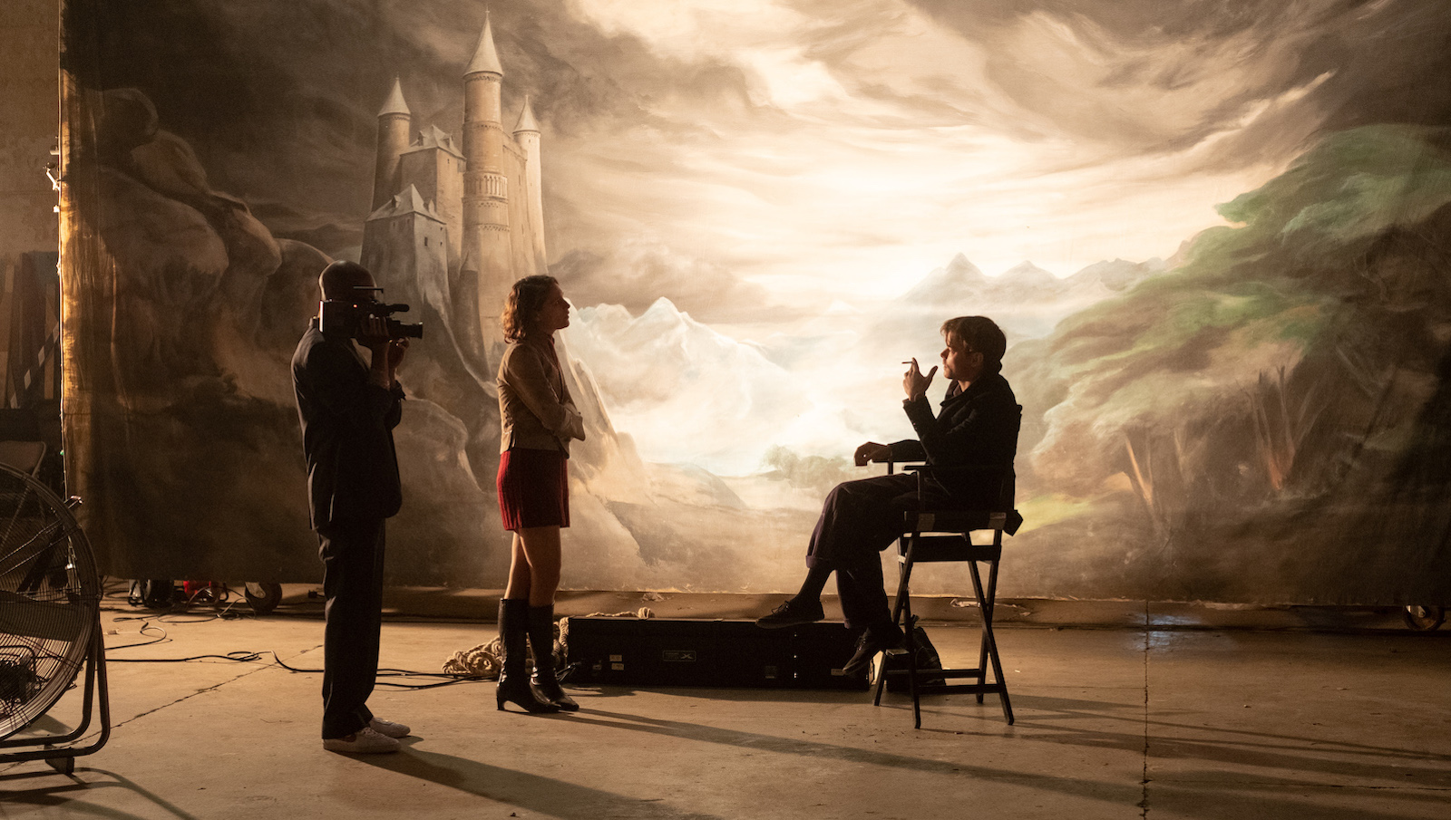 Three people in silhouette, one sitting in a director's chair, on a movie set.
