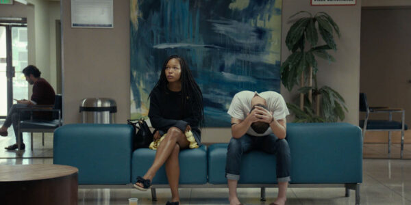 A woman and man sit next to each other in a waiting room; she looks up, his head is buried in his hands.