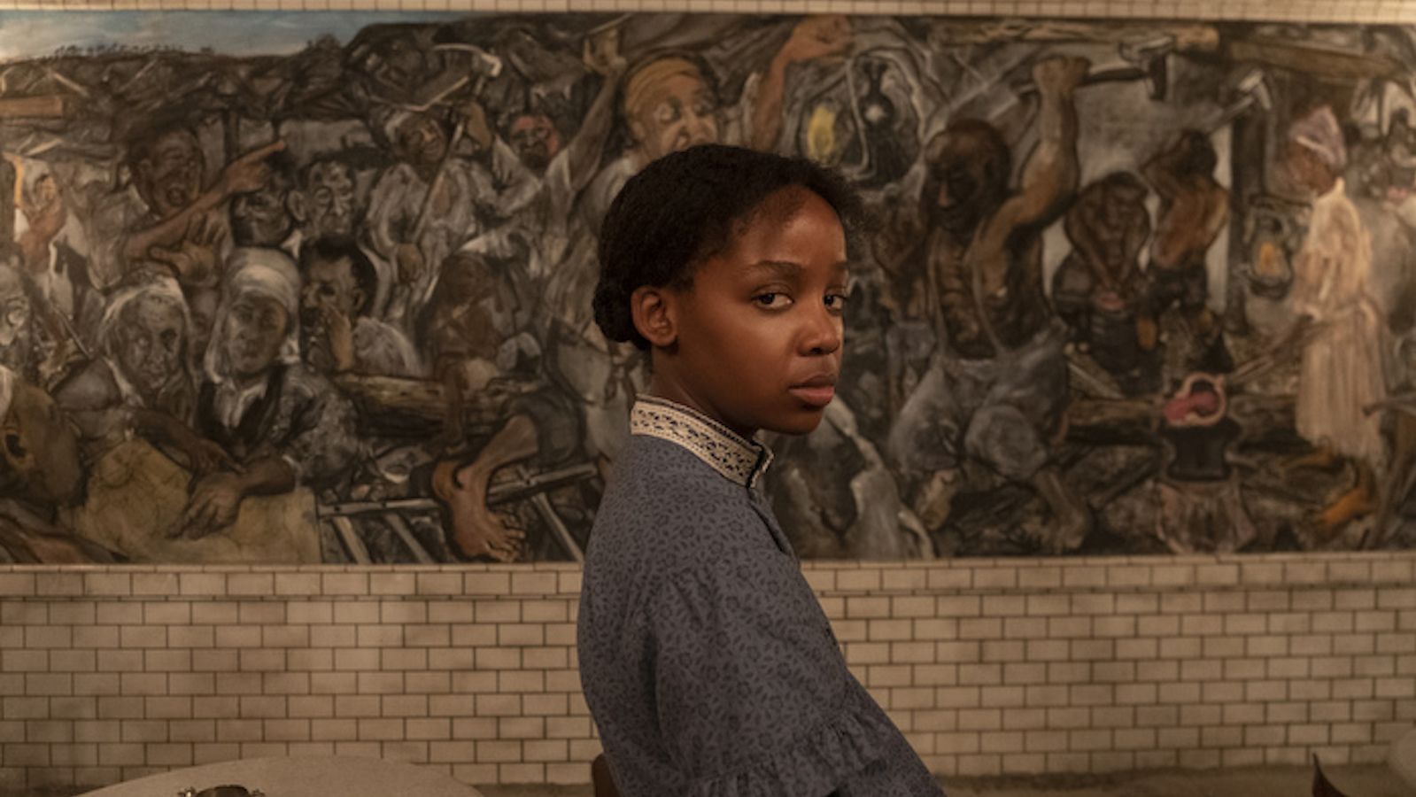 Standing in front of a mural, a young girl in profile turns her gaze on the camera