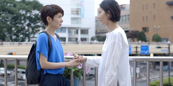 Two women, one in white and one in blue, talk to each other against an outdoor cityscape
