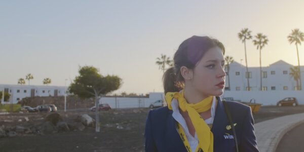 A flight attendant wearing a yellow scarf looks off-camera to the left with a distressed expression, against a sunset.