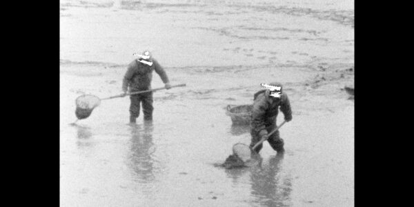 In an aged black and white image, two fisherman stand in knee-deep muddy water with nets, their faces scratched out.