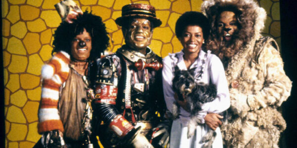 Four cast members of The Wiz, including Dorothy holding her little dog Toto, look at the camera and smile.