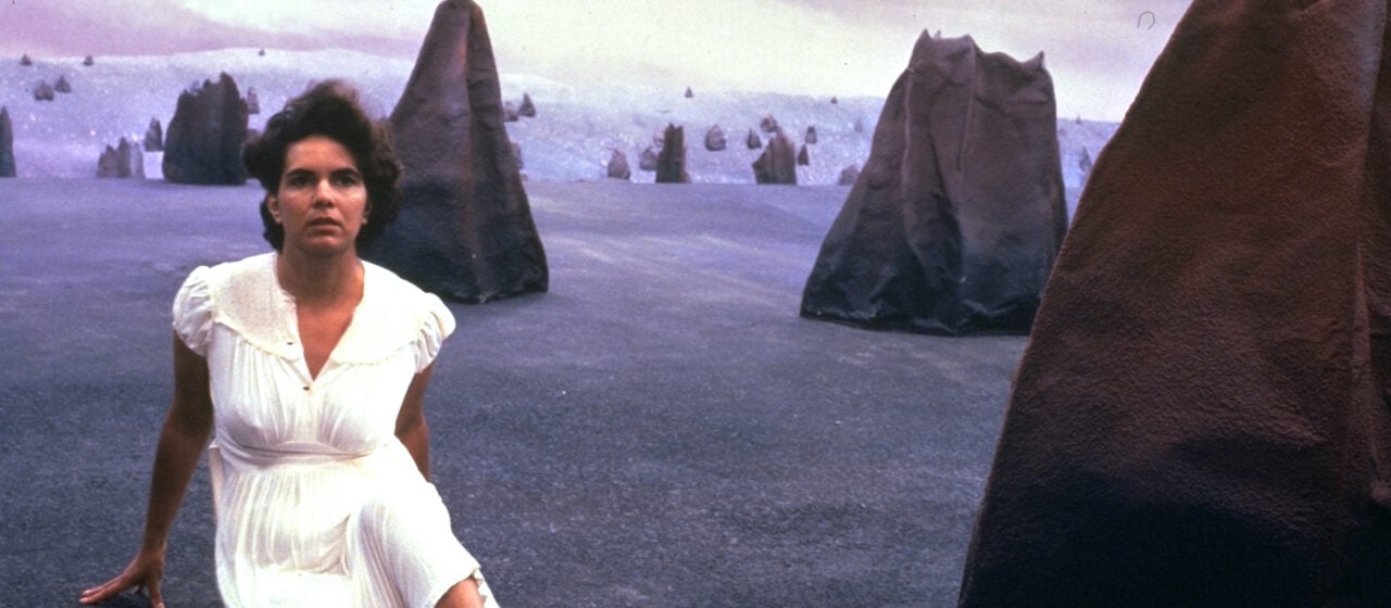 A woman in a white dress sits on a barren landscape surrounded by large, foreboding black rocks