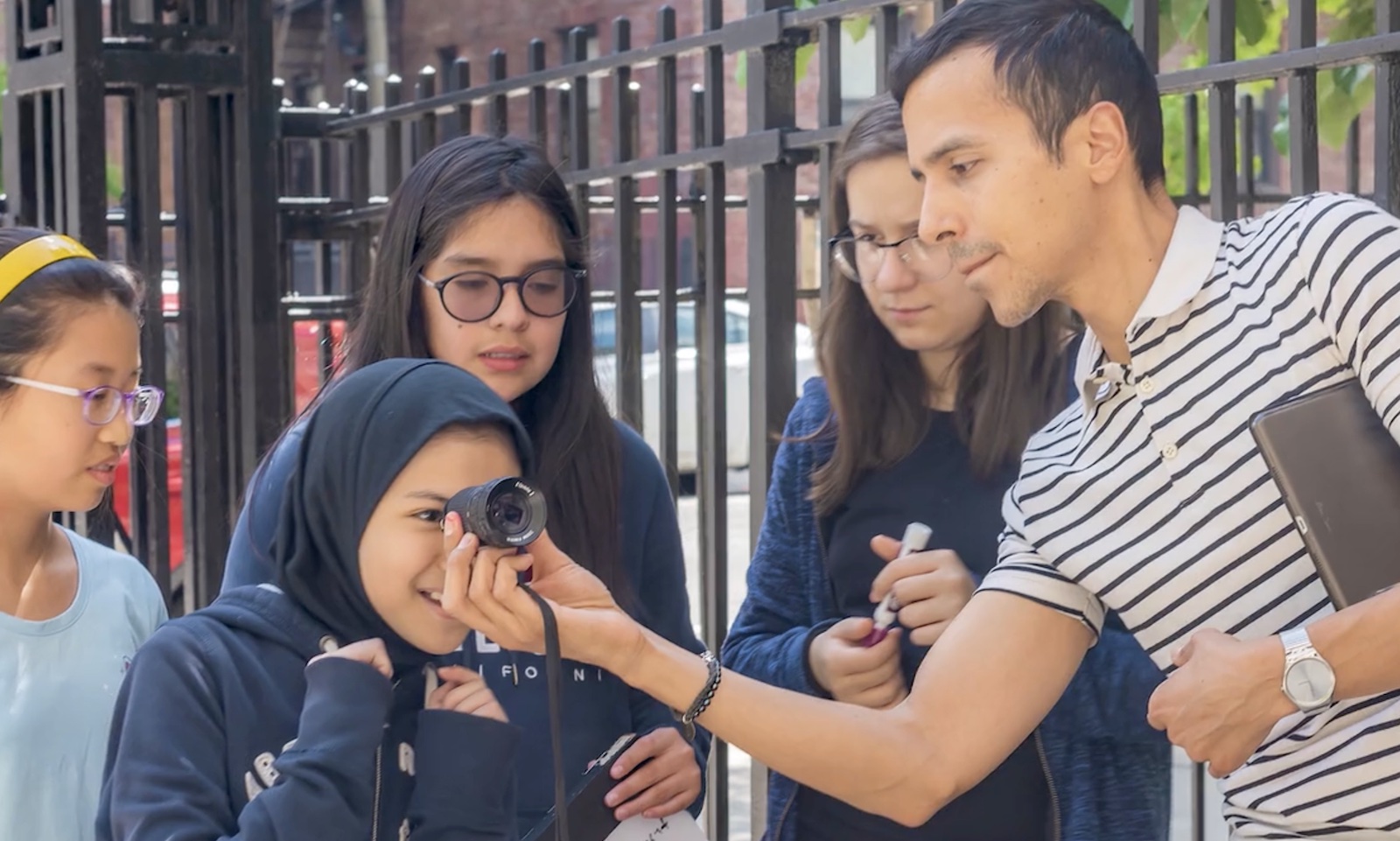 An instructor in a striped shirt holds up a camera lens to the eye of an excited teenage girl, with three teenagers standing behind her