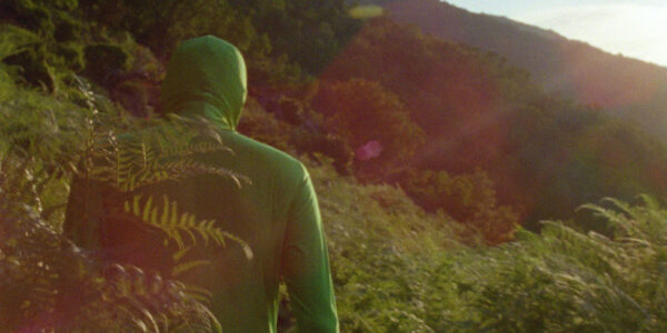 A man dressed entirely in green including over his head is seen from behind looking at a mountainscape