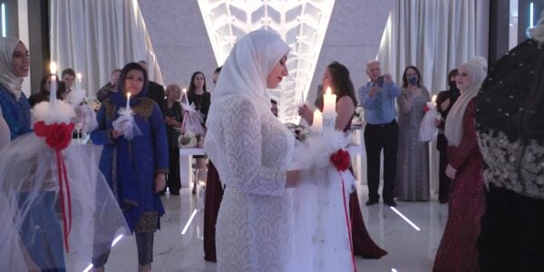 A woman in a white dress and hijab in profile holds a candle in a long hallway with others walking past her
