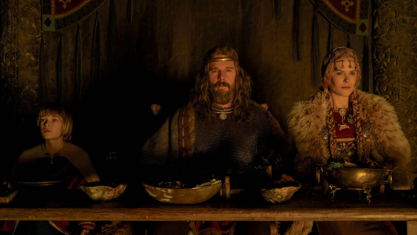 Three members of a Viking family sit in a dark room at a dinner table facing camera