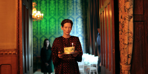 A woman in a hallway holds a birthday cake against a green wallpaper background