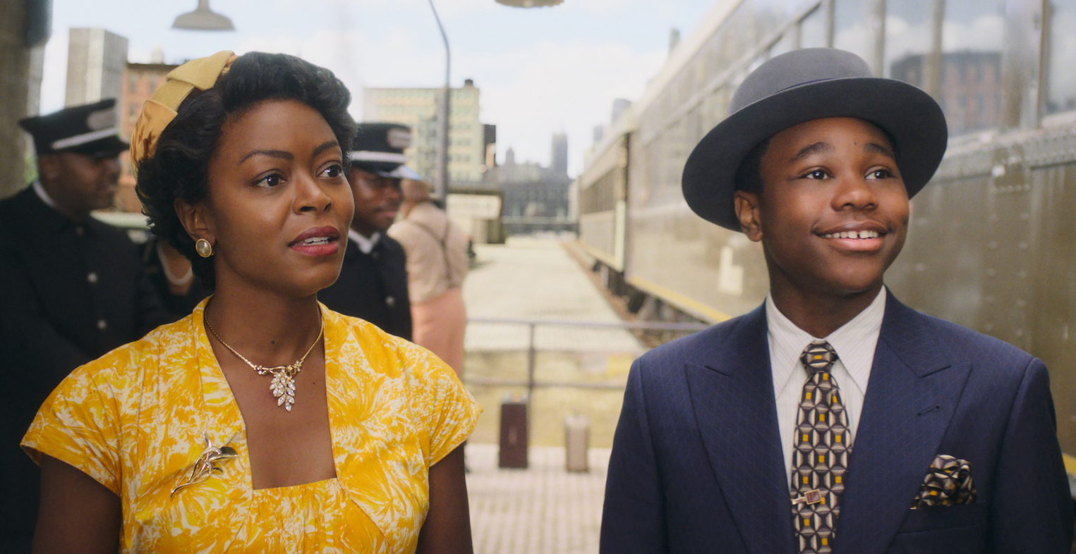 A mother in a yellow dress and her son, in a blue suit and fedora stand on a train platform.