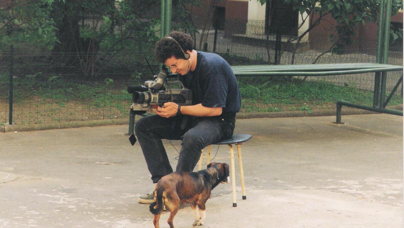 A man sitting in a park looks through a video camera viewfinder while a dog walks by.