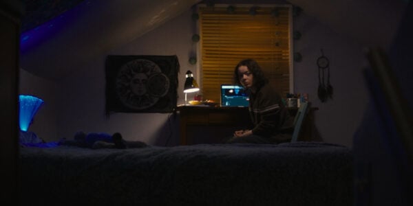 A teenage girl in an attic bedroom looks back at camera from her desk and laptop