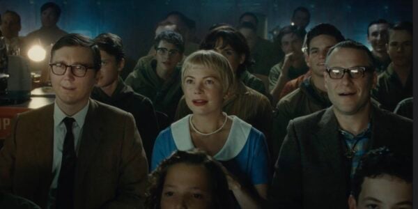 Two men and a woman in the middle in a blue dress look up at a screen with delighted expressions in a dark movie theater