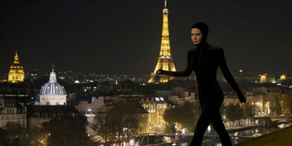 A woman in a tight cat suit walks across a rooftop with the Eiffel Tower behind her, lit up at night