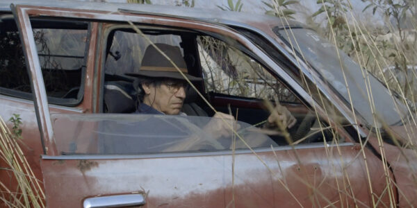 A man sits in a stopped car's passenger seat, the door open, surrounded by tall grass and weeds
