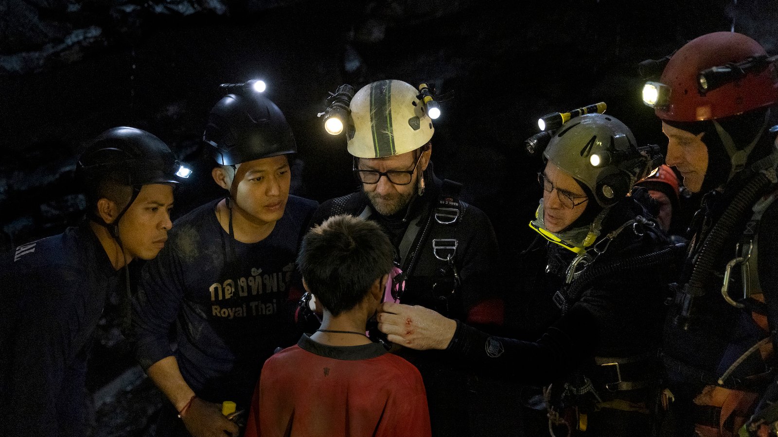 In a cave, a group of divers in helmets stand around a boy, seen from the back, in a red shirt.