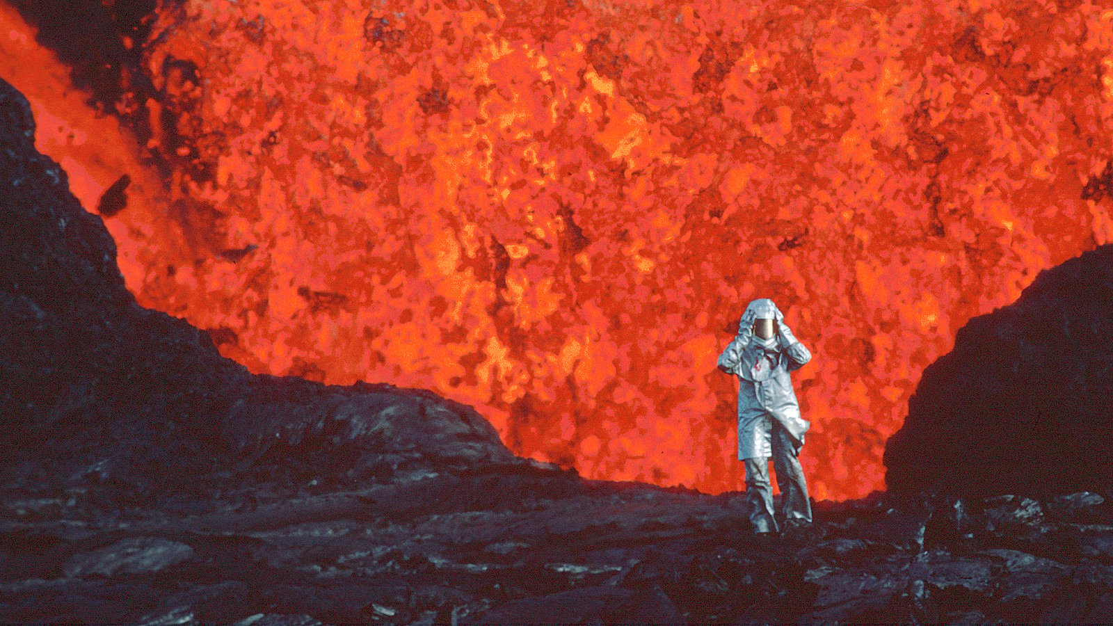 A woman wearing an aluminized suit standing near a red lava burst.